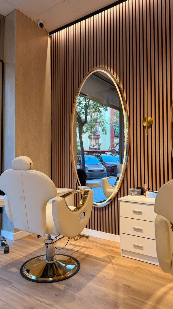 Sillón crema con detalles dorados frente a un espejo ovalado con marco dorado y aplique de pared